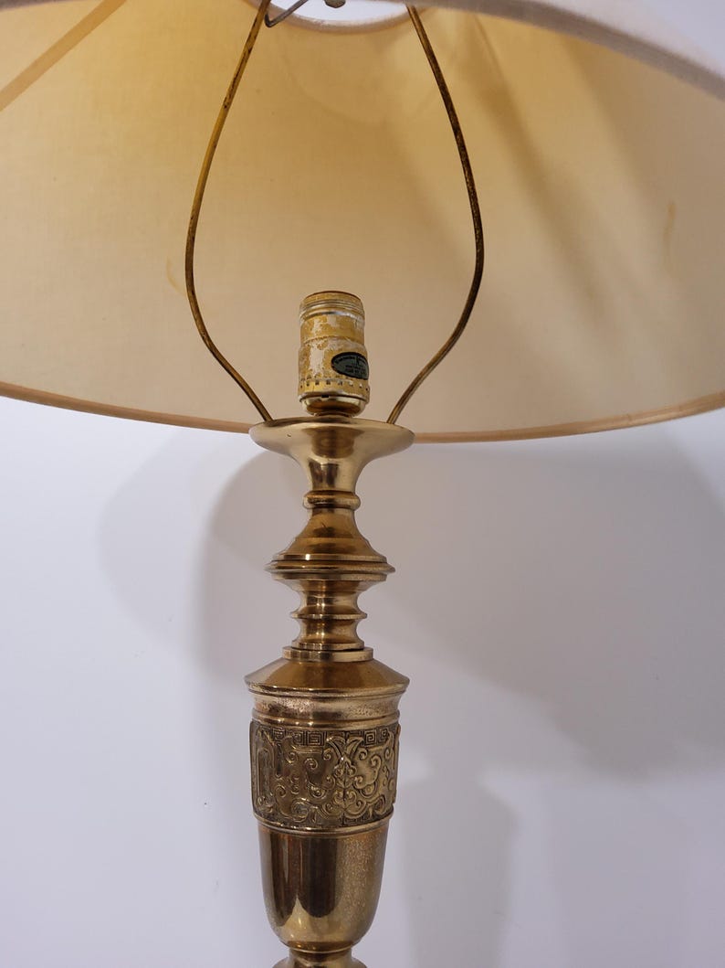 May include: A vintage brass table lamp with a beige lampshade. The lamp features an ornate base with a detailed floral pattern. The lampshade is a soft, neutral color, and the lamp is designed for indoor use.