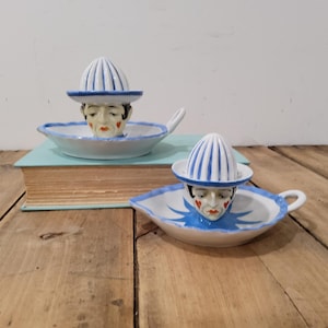 May include: Two ceramic citrus juicers featuring a hand-painted face with red heart-shaped cheeks and lips. Each juicer has a blue and white striped reamer shaped like a hat, a white bowl with a blue scalloped rim, and a small side handle.