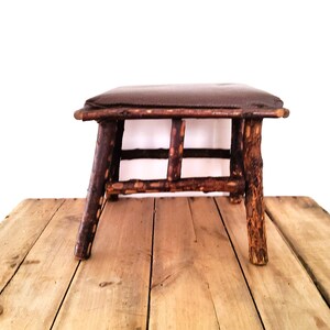 May include: A small stool featuring a dark brown padded seat supported by four legs and horizontal crossbars constructed from natural, unfinished tree branches with a textured, bark-covered surface and a warm, reddish-brown wood tone.