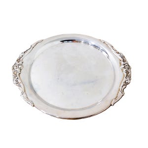May include: A round silver-toned serving tray with a smooth, reflective center surface and a wide, raised rim. The edges of the tray feature intricate, embossed floral and scrollwork detailing on two opposing sides of the circular border.