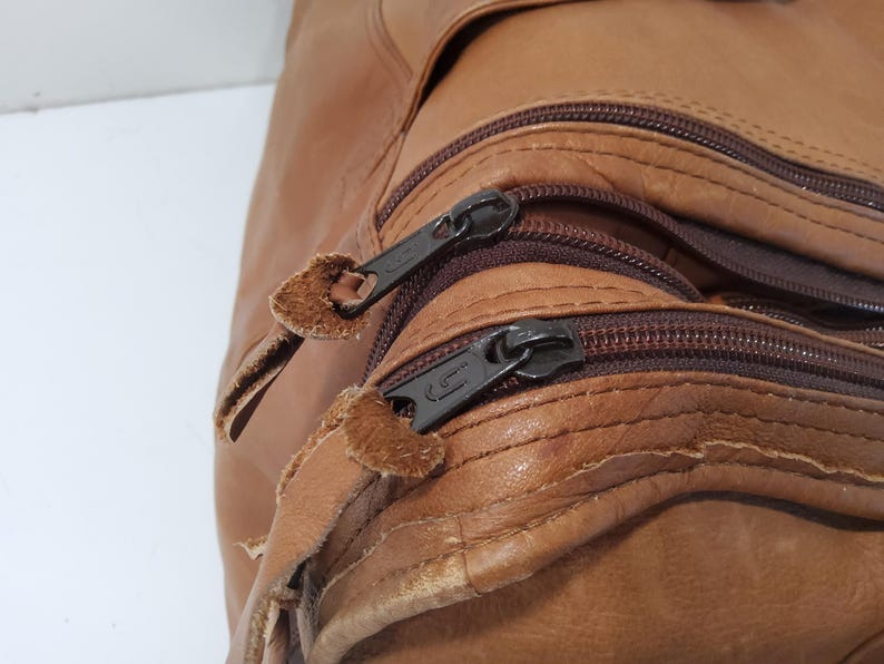 May include: Close-up of a brown leather bag with multiple zippered compartments. The zippers are dark brown with black metal pulls. The leather has a textured appearance.