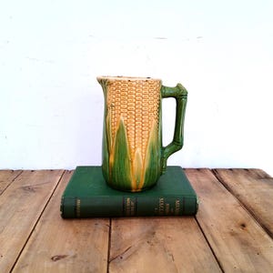 May include: A ceramic pitcher shaped like a corn cob with a yellow kernel texture and green husk leaves at the base. The handle is designed to resemble a stalk. The pitcher is resting on a dark green hardcover book with visible text.