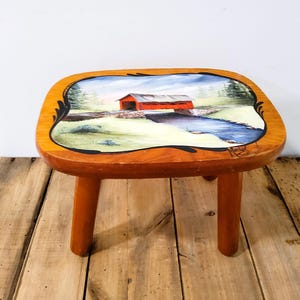 May include: Wooden stool with a hand-painted scene of a red covered bridge over a blue river. The stool has a square top with rounded edges and three wooden legs. The top is painted with a landscape scene.