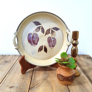 May include: A ceramic serving platter with a beige background and a hand-painted plum design. The platter has two handles and is displayed on a wooden stand. A small terracotta pot with a green plant and a gold candlestick are also visible.