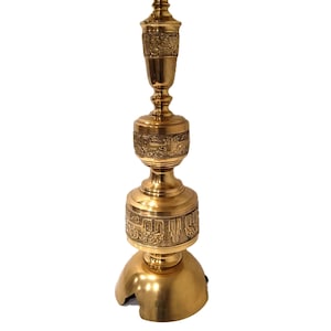 May include: A gold-colored, ornate lamp with a detailed, engraved design. The lamp has a round base, a long, decorative stem, and a top with a flat surface. The lamp is made of metal and has a polished finish.
