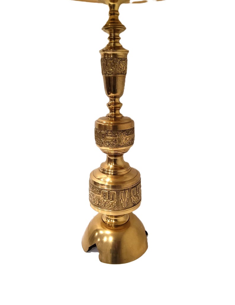 May include: A gold-colored, ornate lamp with a detailed, engraved design. The lamp has a round base, a long, decorative stem, and a top with a flat surface. The lamp is made of metal and has a polished finish.