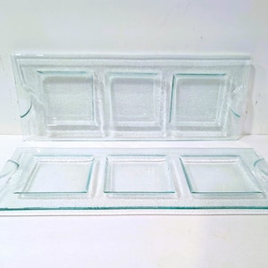 May include: Two clear glass serving trays. Each tray has three square compartments and curved handles. The glass has a textured surface. The trays are rectangular and sit on a white surface.
