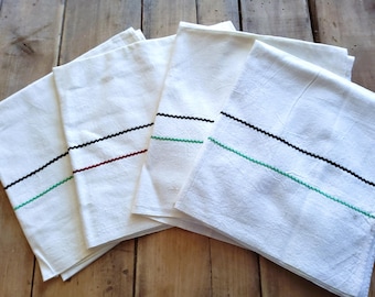 May include: A stack of five white linen kitchen towels with a decorative, zig-zag stitched border. Each towel has a different color border: black, red, and green. The towels are folded and stacked on a wooden surface.