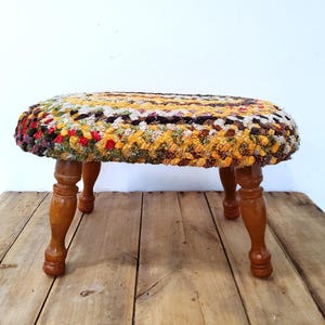 May include: A small, handcrafted stool with a colorful, textured top. The seat is a mix of yellow, red, green, and brown yarns. It has four turned wooden legs, adding a touch of rustic charm. The stool is a unique piece of home decor.