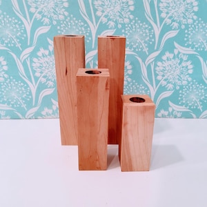 May include: Four unfinished wooden vases of varying heights. The vases are rectangular with a circular opening at the top. The wood has a light brown color. The background is a light blue floral pattern.