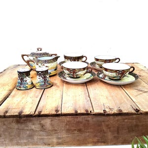 May include: A ceramic tea set featuring a teapot with a lid, a creamer, two salt and pepper shakers, and three teacups with matching saucers. Each piece is decorated with a detailed landscape scene in shades of brown, green, and yellow.