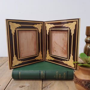 May include: Two wooden picture frames with a geometric design in brown and tan. The frames are empty, with a light wood interior. A small, decorative vase with a plant and a brass candlestick are also visible.