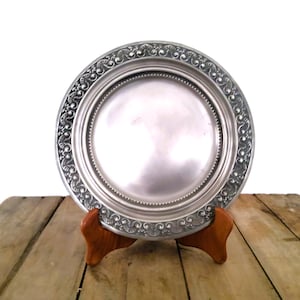 May include: A silver-colored plate with an ornate, embossed border, displayed on a wooden stand. The plate has a reflective surface and a detailed, patterned edge. The wooden stand is a warm brown color.