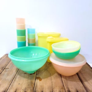 May include: A collection of vintage plastic kitchenware in various colors. Includes stacked cups in peach, green, and blue, along with bowls and containers in green, yellow, and peach. The items are arranged on a wooden surface.