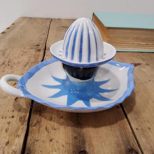 May include: A white ceramic citrus juicer featuring a ribbed dome top with blue stripes. The base is shaped like a leaf with a blue painted sunburst design in the center and a small handle on the side. The piece has a glossy finish throughout.
