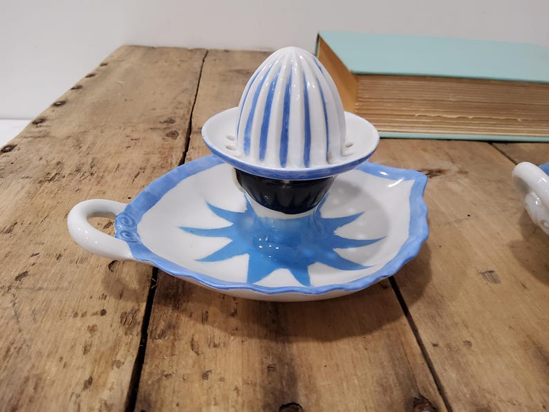 May include: A white ceramic citrus juicer featuring a ribbed dome top with blue stripes. The base is shaped like a leaf with a blue painted sunburst design in the center and a small handle on the side. The piece has a glossy finish throughout.