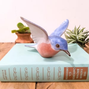 May include: A ceramic bluebird figurine with outstretched wings, featuring a light blue body, white and blue wings, and a reddish-pink breast. The bird is perched on a light blue book with gold lettering. Decorative plants are in the background.