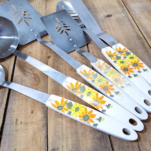 May include: A selection of vintage kitchen utensils, including a ladle, slotted spatulas, and spoons. The handles are white with a painted sunflower design in yellow and orange. The utensils are made of stainless steel.