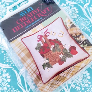 May include: An Avon Creative Needlecraft crewel embroidery kit. The kit features a pillow design with a basket of strawberries, red ribbon, and white flowers. The pillow is framed with a red border and is packaged in a clear plastic bag.