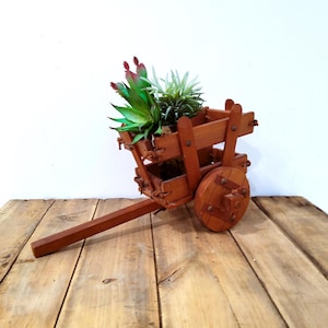 May include: A small, wooden decorative cart planter filled with artificial succulents. The cart is made of stained wood and has a long handle. The cart's wheels are round and attached with metal studs. The cart is sitting on a wooden surface.