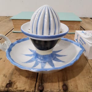 May include: A white ceramic citrus juicer featuring a ribbed dome top, a scalloped edge, and a blue painted starburst pattern on the base. The juicer includes a curved handle with a spiral detail and a central dark blue band around the reamer.