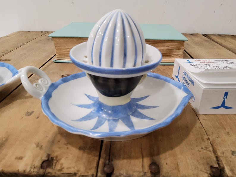 May include: A white ceramic citrus juicer featuring a ribbed dome top, a scalloped edge, and a blue painted starburst pattern on the base. The juicer includes a curved handle with a spiral detail and a central dark blue band around the reamer.