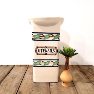 May include: Cream-colored ceramic utensil holder with a rectangular shape. It features a decorative band with green leaves and a brown and white design. The word "UTENSILS" is displayed in a brown, decorative frame. A small wooden vase with a green plant is next to it.