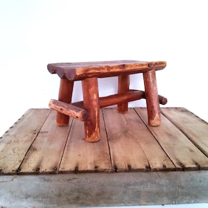 May include: A small, low-profile bench constructed from reddish-brown, unfinished logs. The seat is made of three parallel logs, supported by four cylindrical log legs and reinforced with horizontal log stretchers connecting the legs on each side.