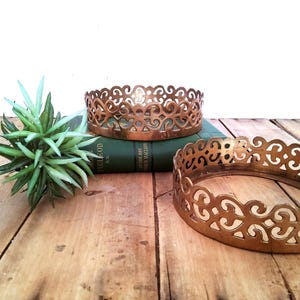 May include: Two decorative, openwork metal rings with a scrolled design in a warm gold tone. One ring is on top of a green book, and the other is on a wooden surface. A small green plant is also on the wooden surface.