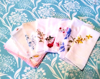 May include: A collection of white handkerchiefs with embroidered floral designs, including cherries, blue and yellow flowers, and delicate branches. The handkerchiefs are arranged on a turquoise patterned surface.