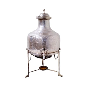 May include: Silver-colored, ornate water dispenser with a floral pattern. The dispenser has a spigot, a lid, and sits on a decorative stand with three legs. The stand has chains attached to the dispenser.
