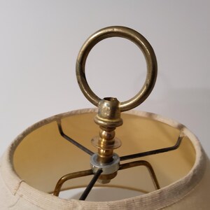 May include: Close-up of a vintage lamp shade with a brass ring and metal frame. The shade is a light beige color, and the metal components have a warm, aged patina. The lamp appears to be an antique or vintage style.