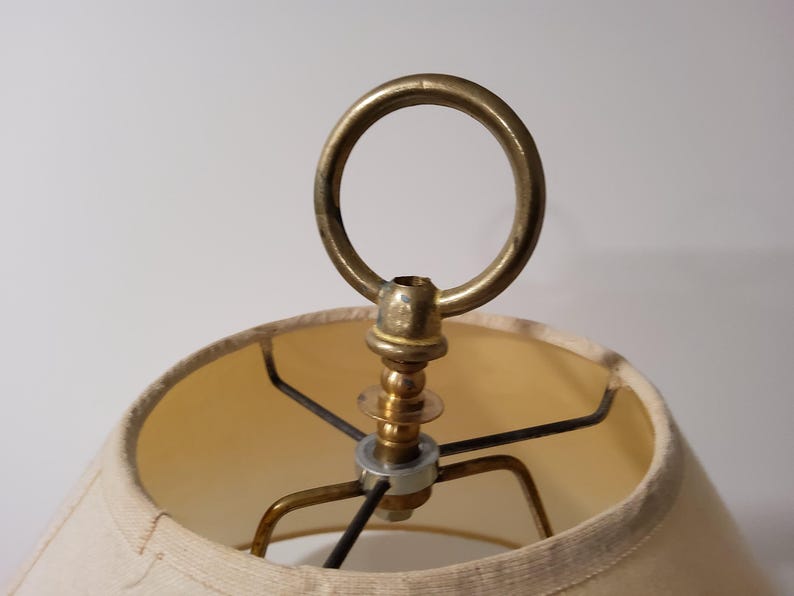 May include: Close-up of a vintage lamp shade with a brass ring and metal frame. The shade is a light beige color, and the metal components have a warm, aged patina. The lamp appears to be an antique or vintage style.
