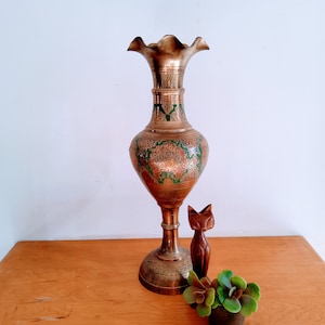 May include: A tall, ornate brass vase with green and gold floral designs. The vase has a wide mouth and a flared base. It is sitting on a wooden table with a wooden cat figurine and a potted succulent plant.