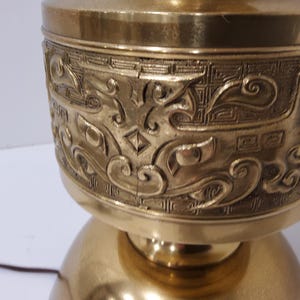 May include: A brass-colored lamp with an ornate, engraved design. The lamp features a detailed pattern of swirling lines and geometric shapes around the upper portion. The base is a rounded shape, and the lamp has a warm, metallic finish.