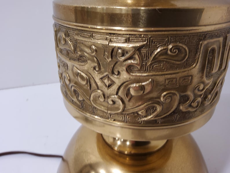May include: A brass-colored lamp with an ornate, engraved design. The lamp features a detailed pattern of swirling lines and geometric shapes around the upper portion. The base is a rounded shape, and the lamp has a warm, metallic finish.