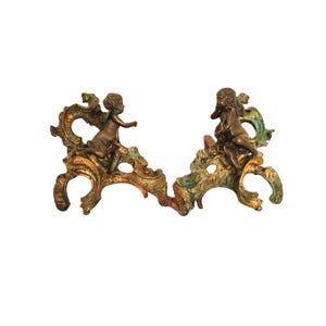 May include: A pair of bronze fireplace andirons featuring two cherub figures perched atop ornate, scrolling metal bases. The metal has a weathered, multi-toned finish with dark brown, gold, and verdigris green accents throughout the intricate design.