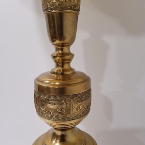 May include: A tall, ornate brass candlestick with intricate engraved patterns. The candlestick features a flared base, a central bulbous section, and a slender top, all adorned with detailed relief designs. The overall design is symmetrical and the brass has a polished finish.