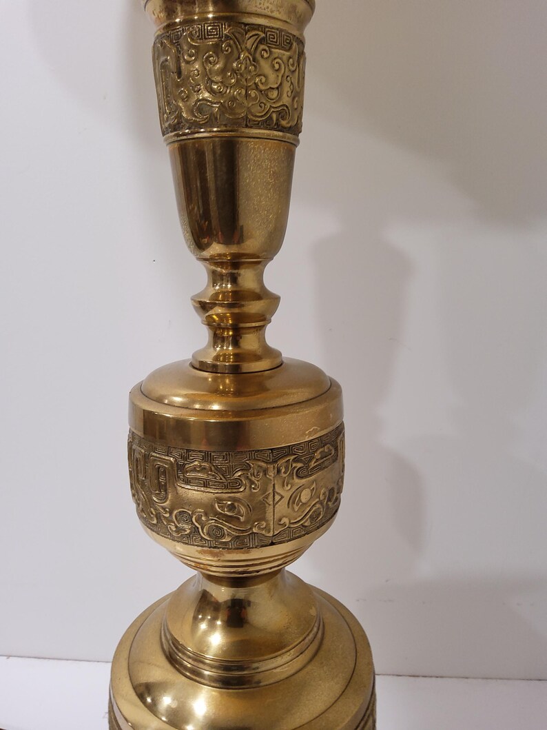 May include: A tall, ornate brass candlestick with intricate engraved patterns. The candlestick features a flared base, a central bulbous section, and a slender top, all adorned with detailed relief designs. The overall design is symmetrical and the brass has a polished finish.
