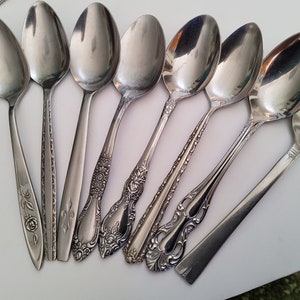 May include: A set of nine silver spoons with ornate designs on the handles. The spoons are arranged in a fan shape on a white surface.