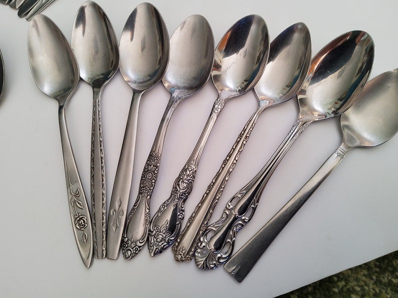 May include: A set of nine silver spoons with ornate designs on the handles. The spoons are arranged in a fan shape on a white surface.
