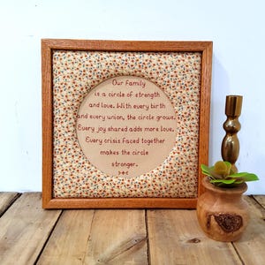 May include: A framed embroidery with the text "Our Family is a circle of strength and love." The frame is wood, and the fabric has a floral pattern. A small wooden pot with a plant and a gold candle holder are next to the frame.