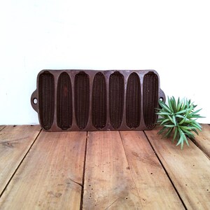 May include: A dark brown cast iron corn stick pan featuring seven individual molds shaped like ears of corn. The rectangular pan has a handle on each side and a textured surface within each mold to create the detailed kernel pattern.