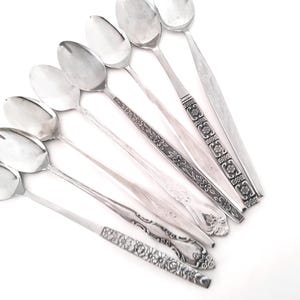 May include: A selection of vintage silver spoons with diverse handle designs, including floral and geometric patterns. The spoons are arranged on a white surface, displaying their polished, reflective surfaces. A collection of silverware.