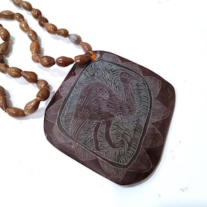 May include: A brown necklace with a large, square pendant. The pendant features an etched design of a bird within a floral border. The necklace has a beaded chain made of small, brown beads.