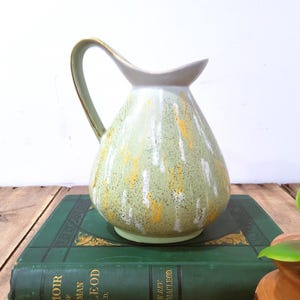 May include: A vintage ceramic pitcher with a light green base and a speckled pattern of white and yellow. The handle and rim are a contrasting light gray with gold trim. The pitcher is displayed on a green book.