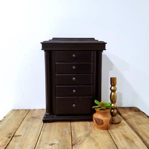 May include: A dark brown wooden jewelry box with five drawers, a small decorative pot with a green plant, and a gold-colored candlestick. The jewelry box is rectangular and has a classic design. The items are on a wooden surface.