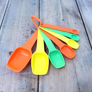 Puede incluir: Un juego de cucharas medidoras coloridas en naranja, amarillo y verde. Las cucharas están dispuestas en forma de abanico, con un anillo en la parte superior para colgar. Las cucharas son de plástico y están sobre una superficie de madera.