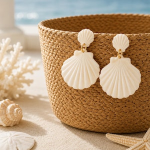 May include: A pair of off-white shell-shaped drop earrings featuring a small fan-shaped stud connected to a larger, ribbed scallop shell pendant by a small gold-toned metal ring. The earrings are displayed hanging over the edge of a woven basket.
