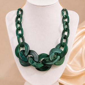 May include: A chunky statement necklace featuring large, interlocking oval links in a deep, marbled forest green resin. The necklace is connected to a thin gold-toned metal chain at the top, creating a bold, circular design around the neck.